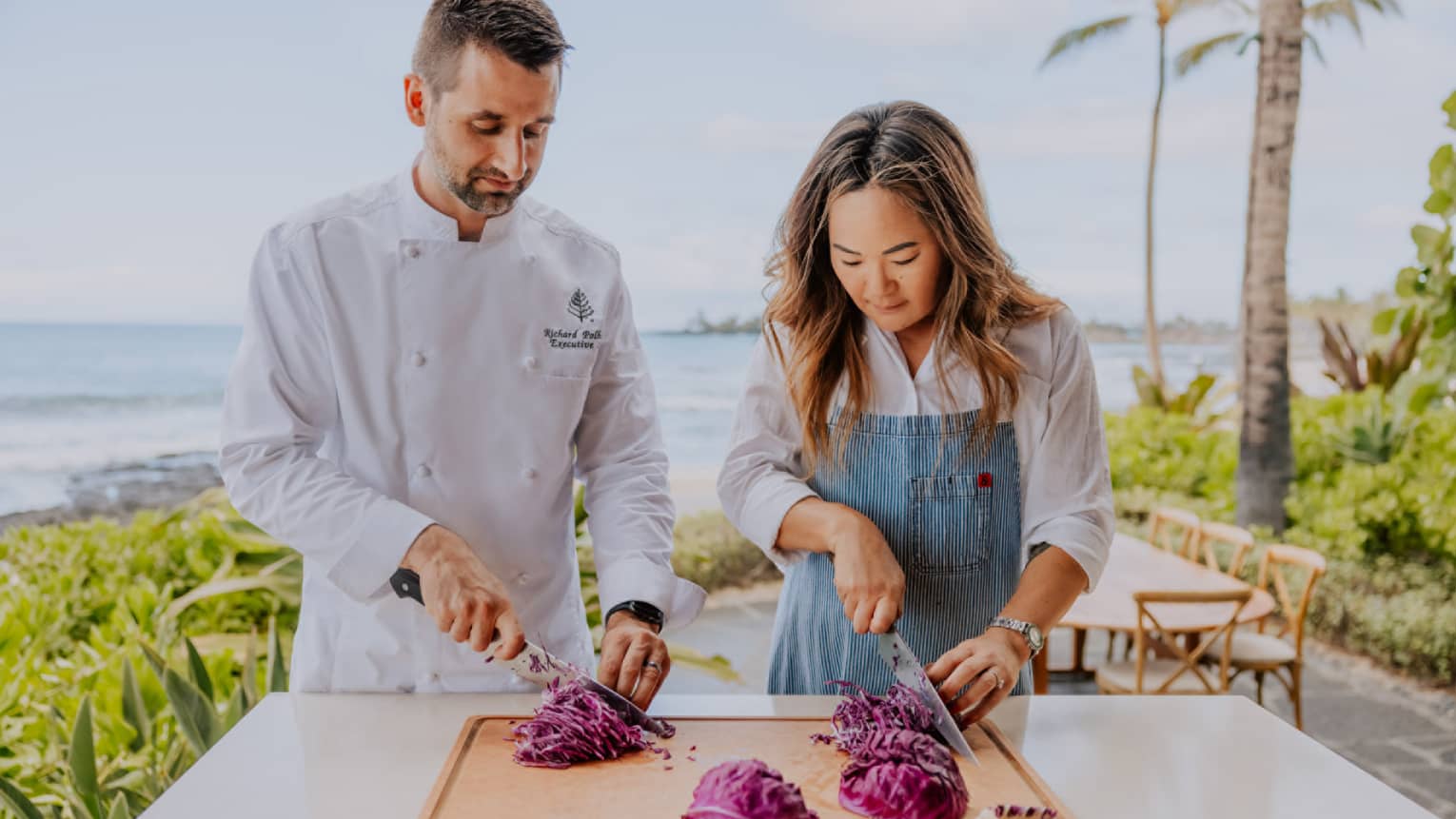 A woman and a man in a chef's jacket practice thinly slicing vegetables