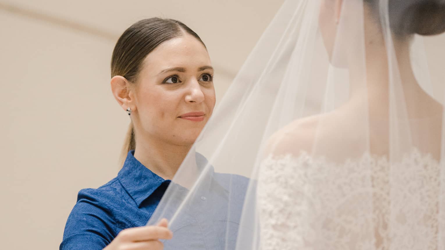 Wedding specialist wearing blue dress facing bride and holding her veil with right hand