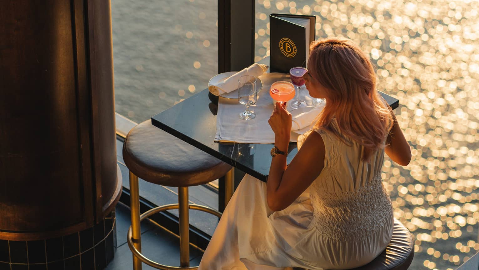 A person wearing a dress and sipping a drink while sitting at a dining table by a window
