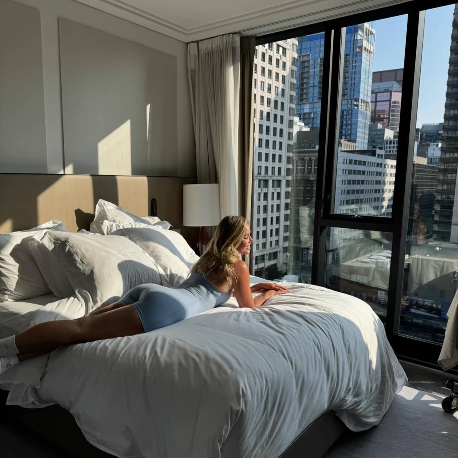 A woman lounges on a plush bed by a large window overlooking a city view