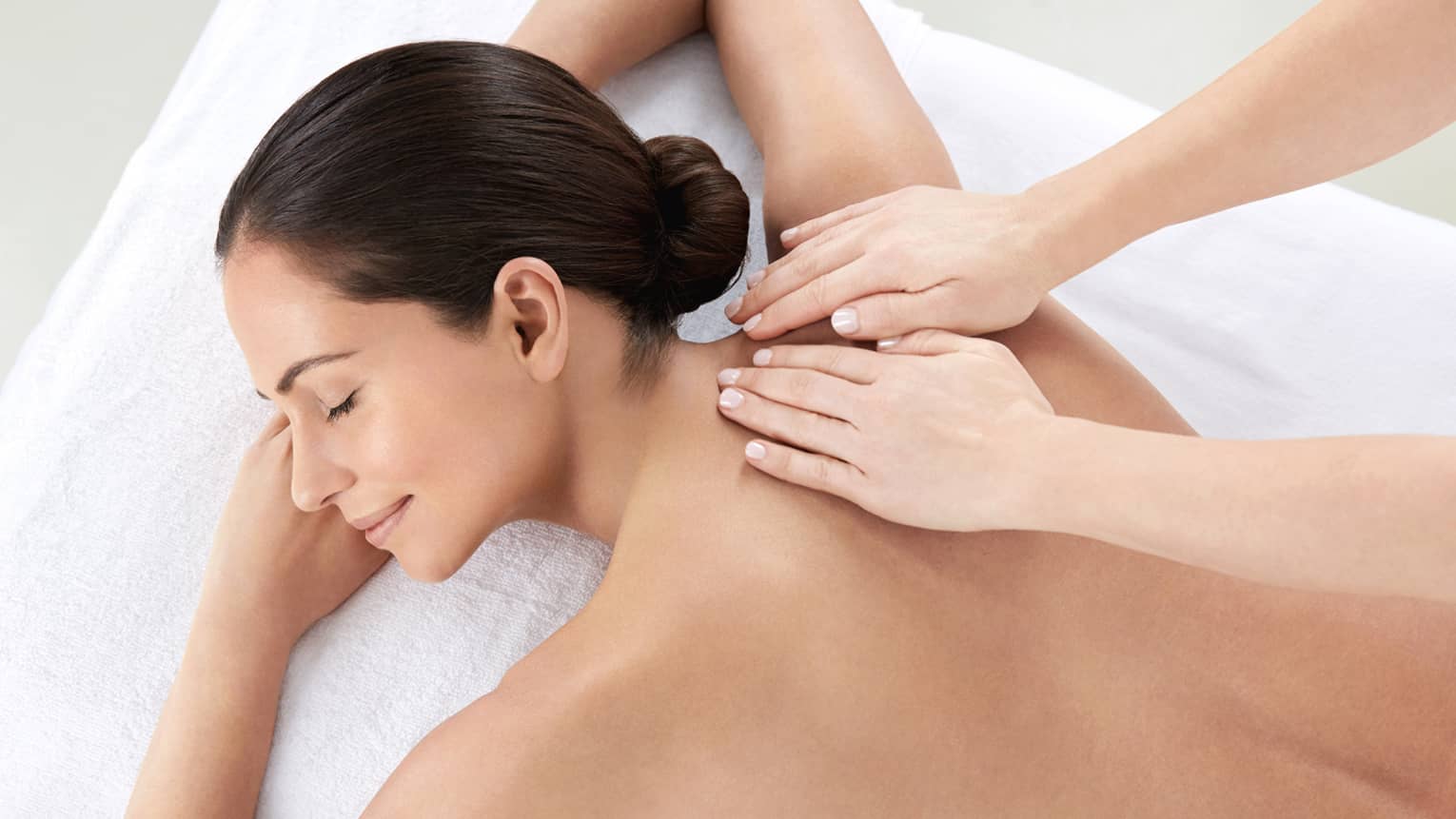 Woman smiles, lies on massage table as hands massage bare shoulders