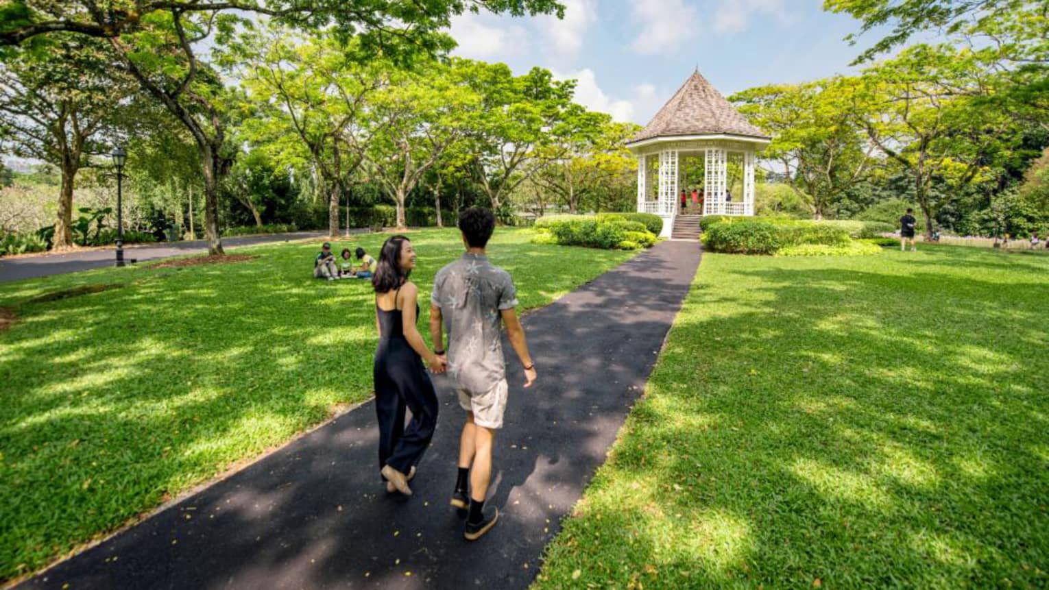 A man and woman hold hands and walk along an asphalt trail through the Singapore Botanic Gardens