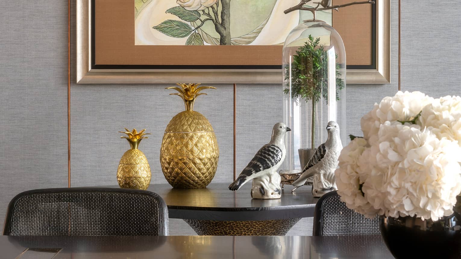 Dining area featuring a wooden table, decorative golden pineapples, ceramic bird figurines and a vase of white flowers. A framed botanical art piece hangs on the wall above.