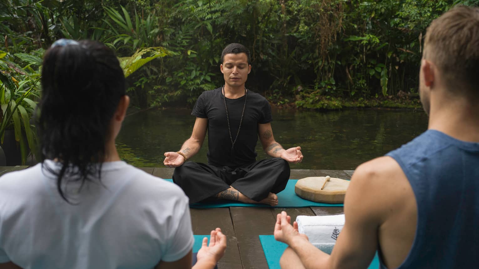 Two people sit with their backs to the camera looking onto an instructor who is sitting cross-legged and with hands in the Gyan mudra position