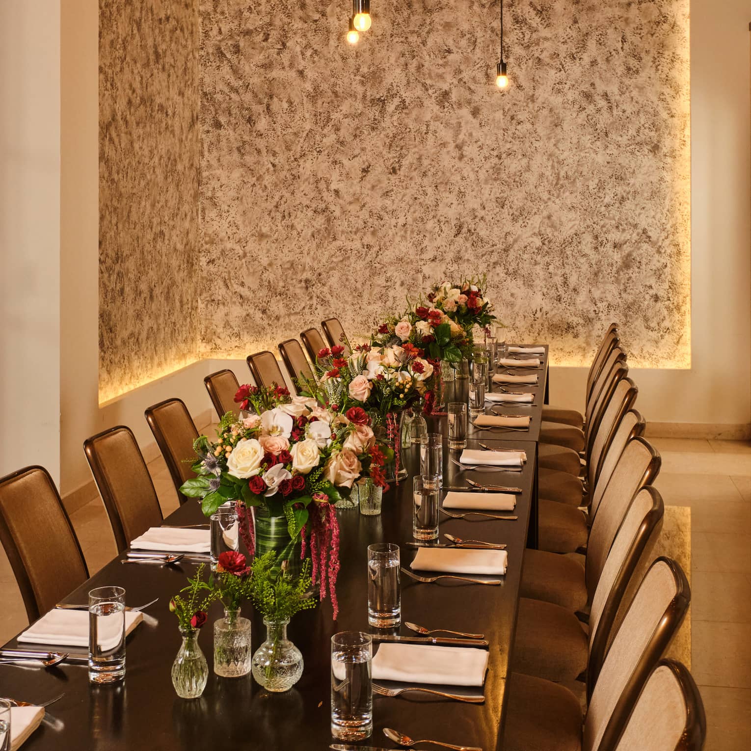 Long rectangular dining table set in a private dining room with red-and-light-pink floral arrangements in the centre and string lights overhead