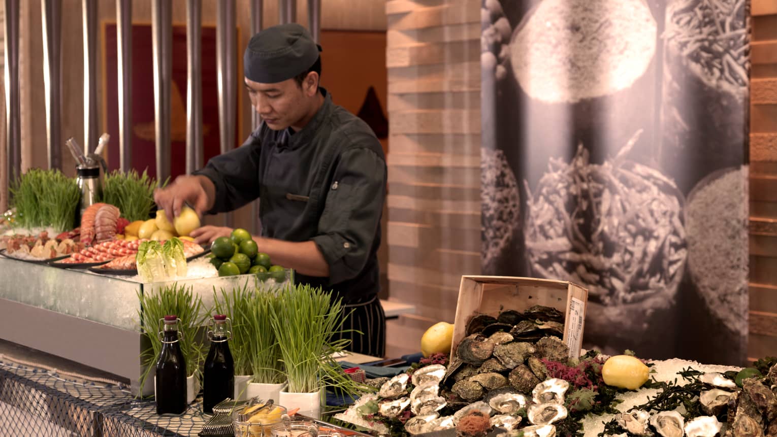 Man in black cook's uniform stands behind buffet with platters of fresh raw seafood on ice, citrus fruit
