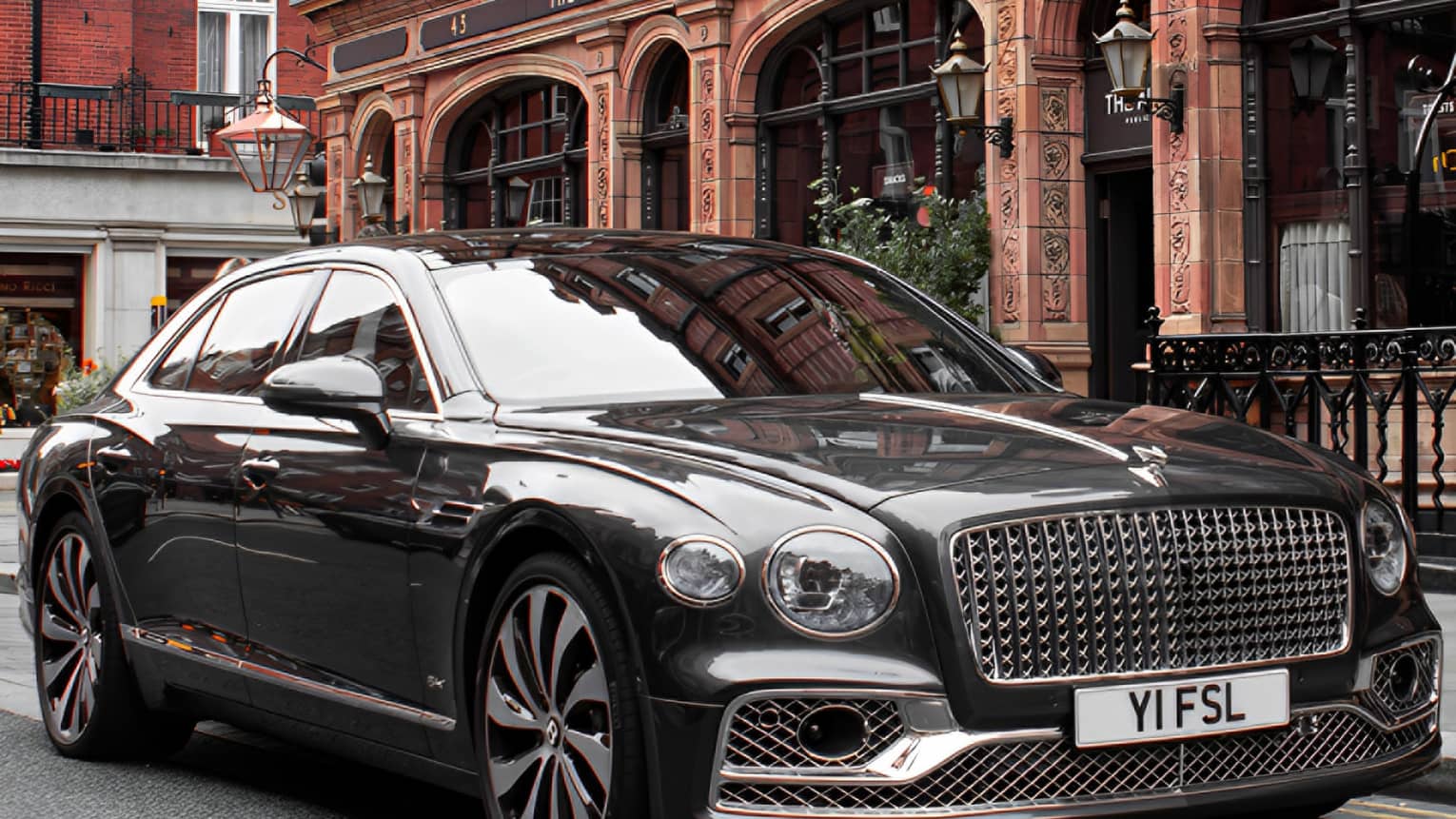 Close-up front view of shiny black modern Bentley with an elaborate grille parked in front of a restored Victorian building.