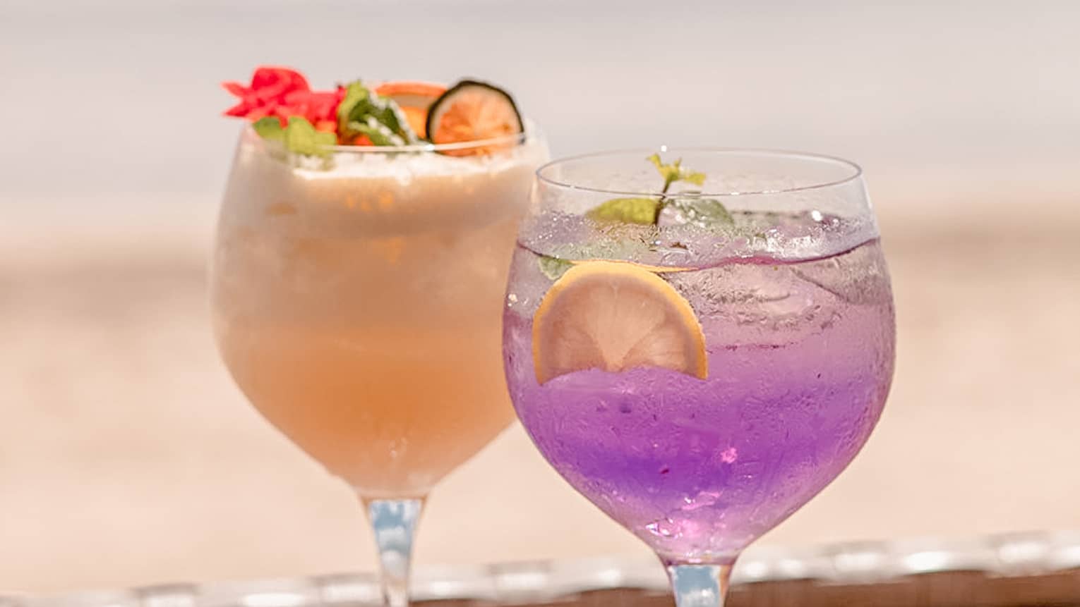 Two colourful cocktails in stemmed glasses on wooden tray, beach in background