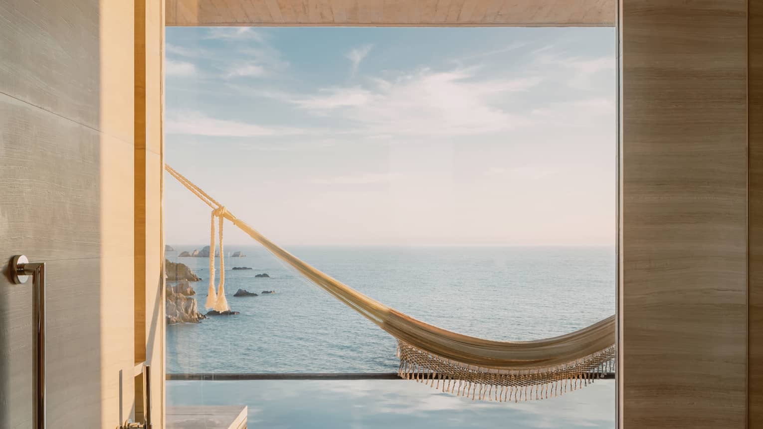 Minimalist bathroom with a deep tub and a large window offering a breathtaking view of an infinity pool, a hammock and the ocean stretching towards the horizon