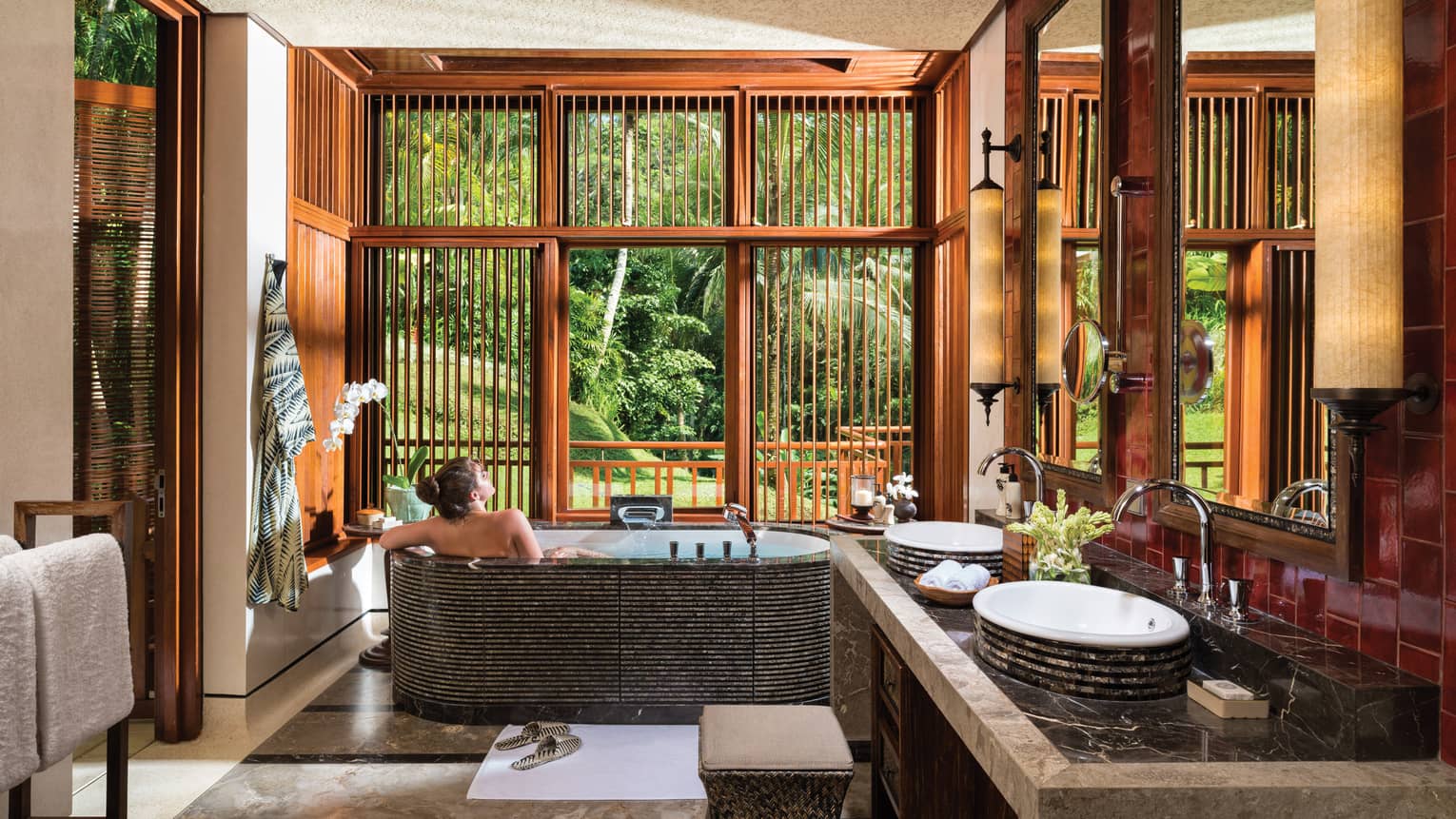 Woman with bare back relaxes in black freestanding spa tub near double sink, wood shutters open to lush garden view