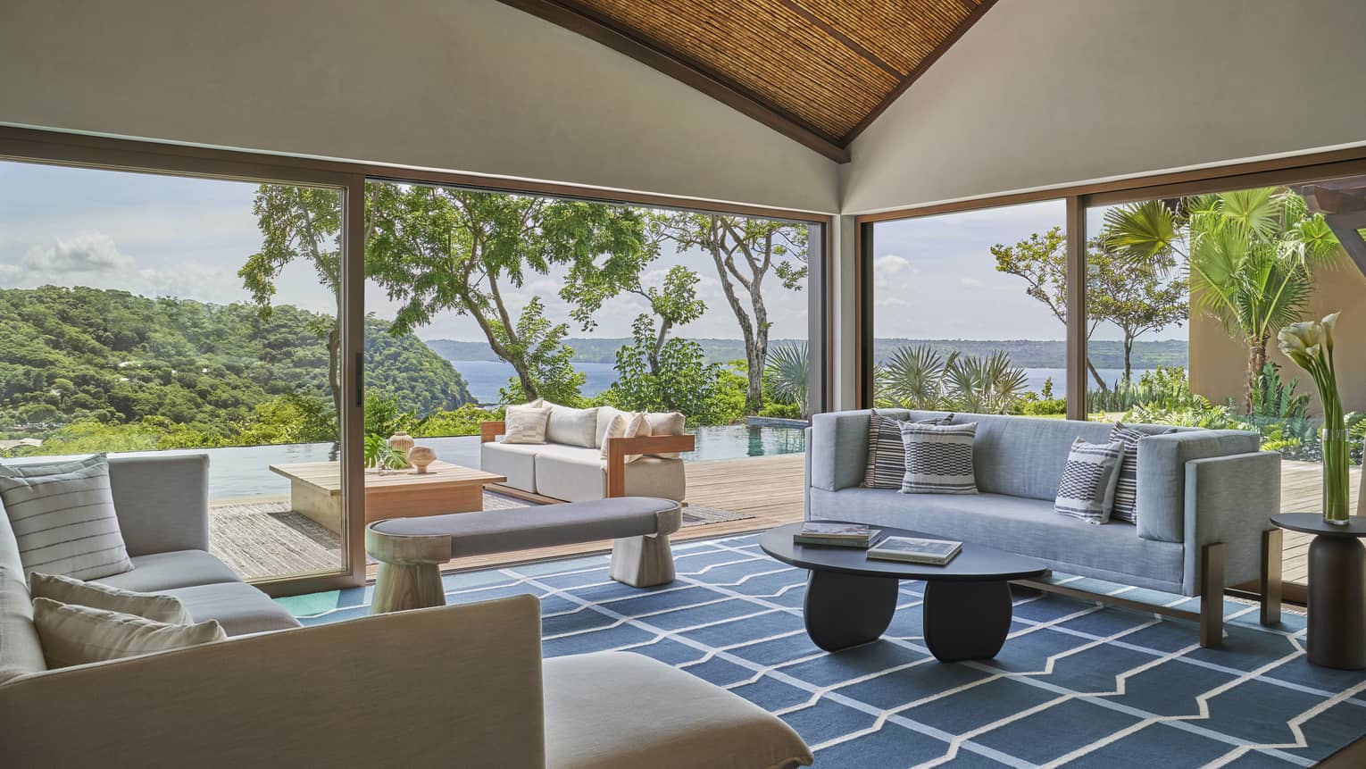 Living room with blue and white rug, two sofas, bench and coffee table, two walls of windows look at to pool area and sea