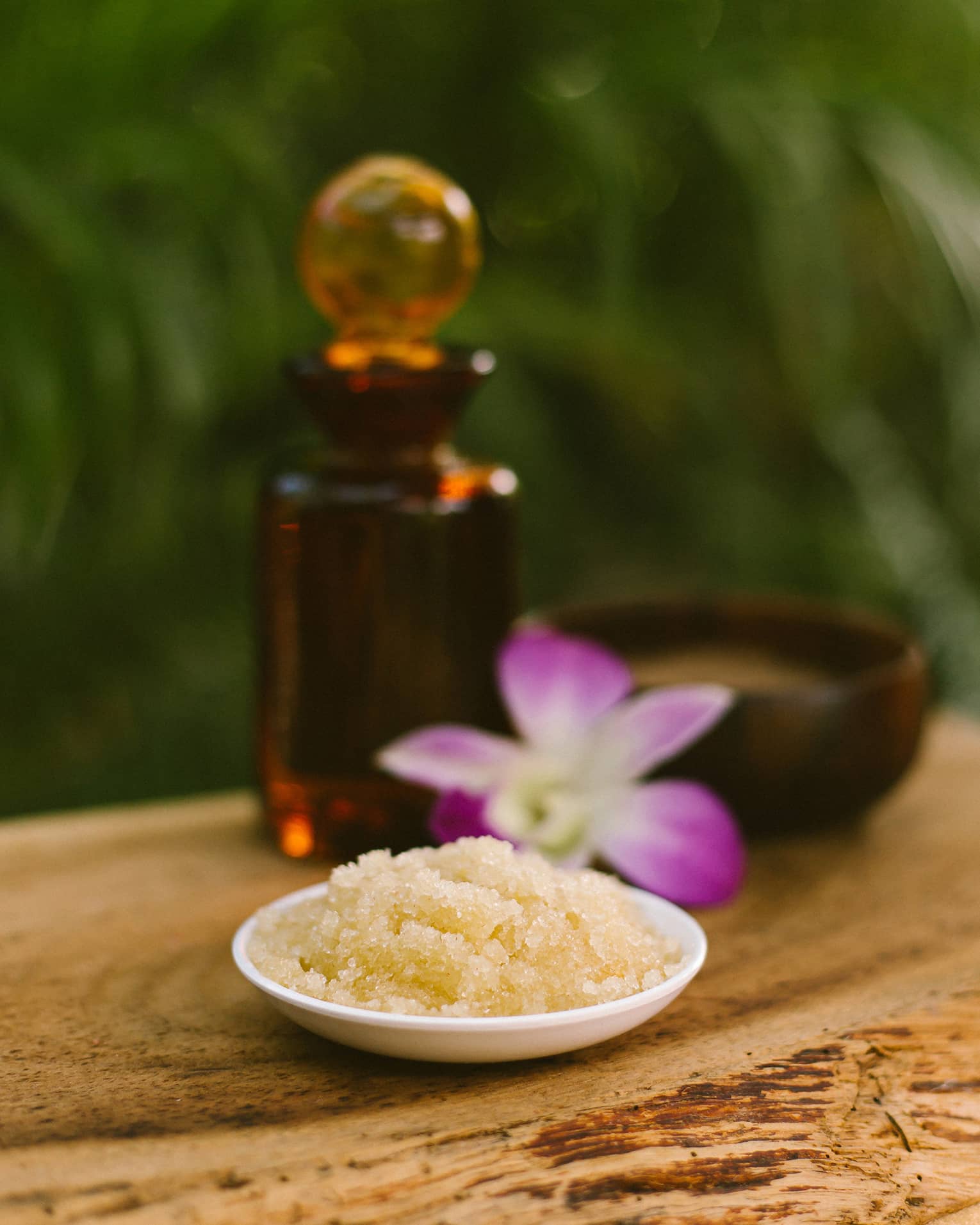 Bowl with salt scrub next to purple tropical flower and glass oil bottle on rustic table