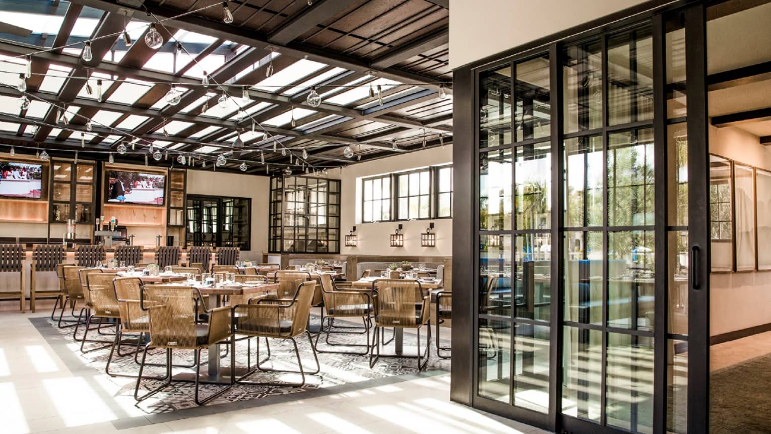 Dining room and bar with glass roof, modern wicker chairs and tables, glass sliding doors with black frames, glass bulb light strings
