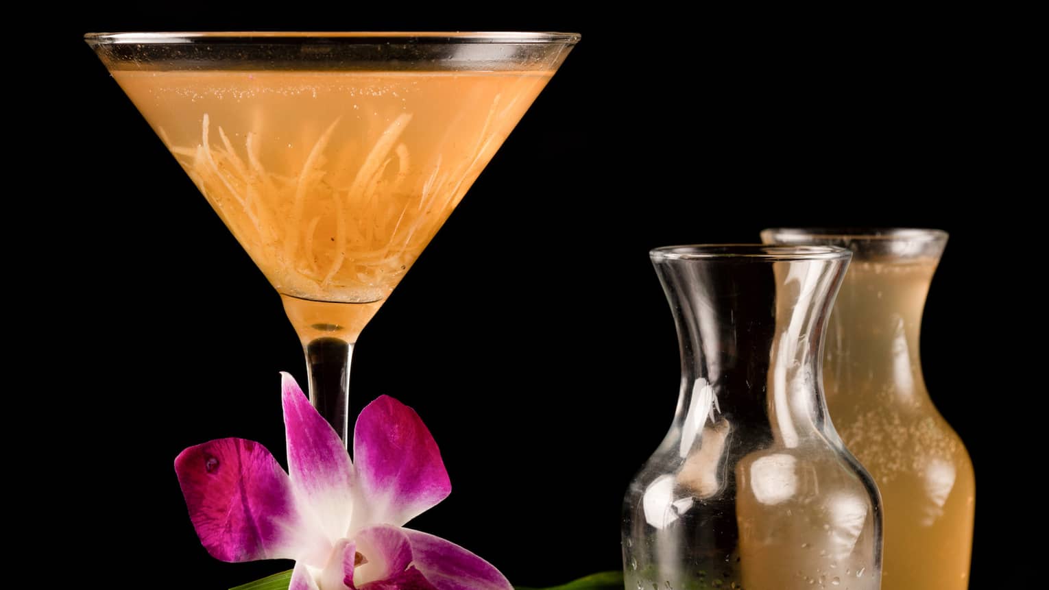 An orange martini glass on a wooden table surrounded by several pitchers of alcohol.