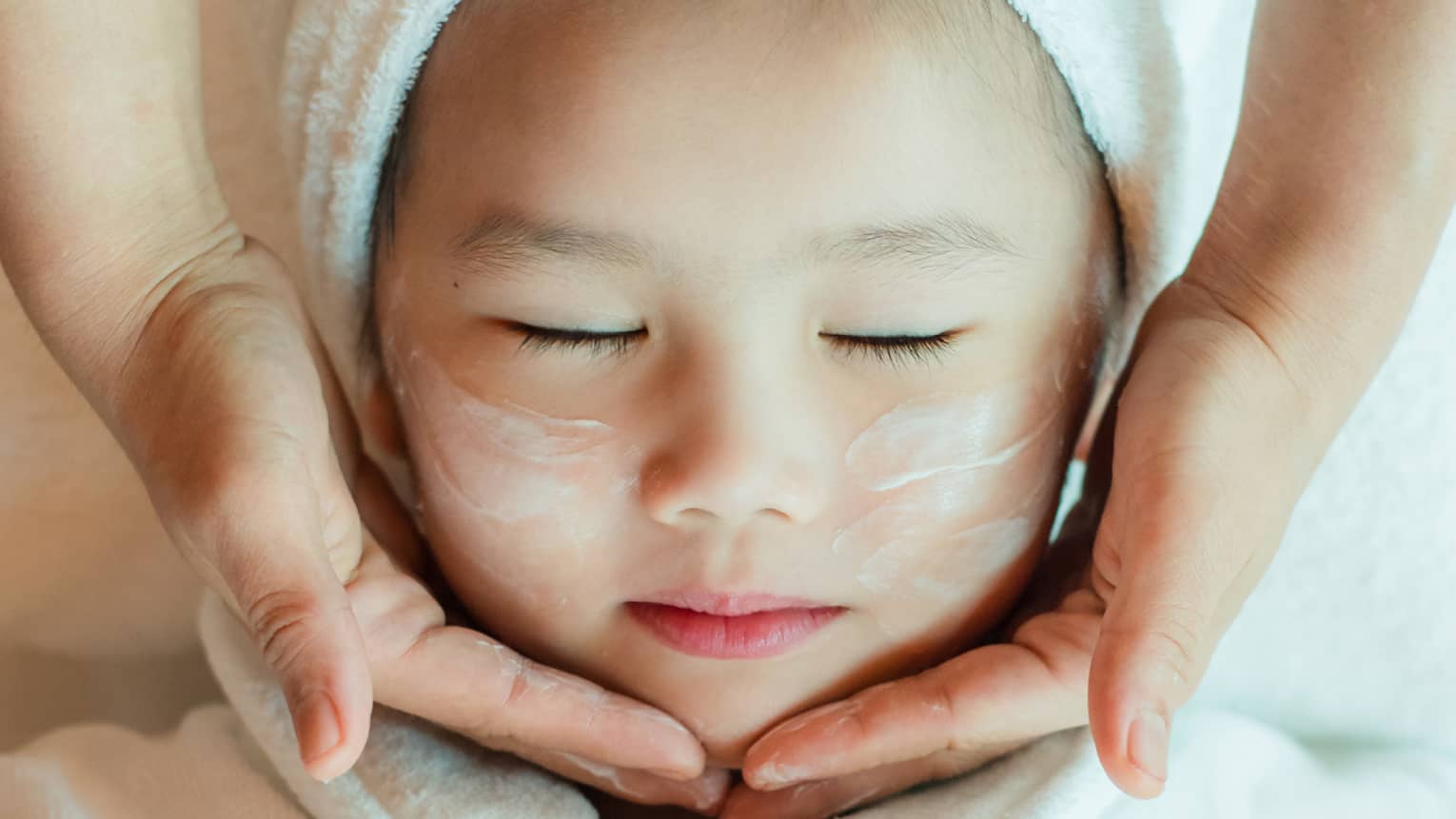 A close-up of a relaxed child wrapped in a towel and robe and enjoying a masseuse massaging a cream into their face.