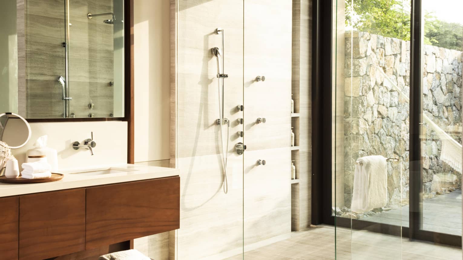 A modern bathroom with a spacious glass-enclosed shower, wooden vanity and large mirror. Wicker stools add texture, while natural light enters through glass doors overlooking an outdoor space.