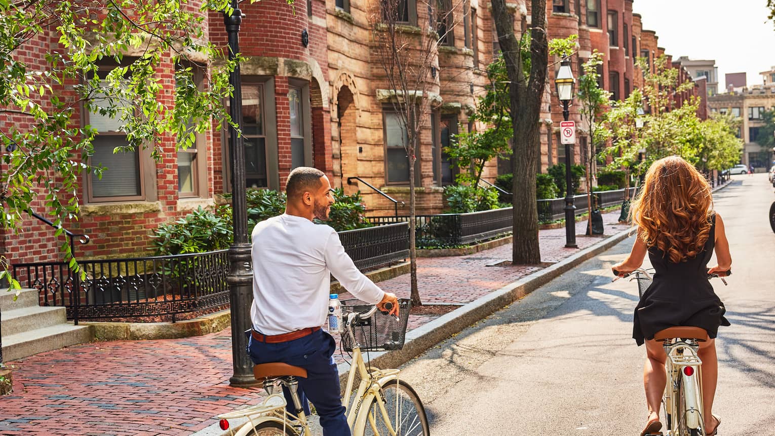 A couple rides bicycles along a street lined with brownstone homes