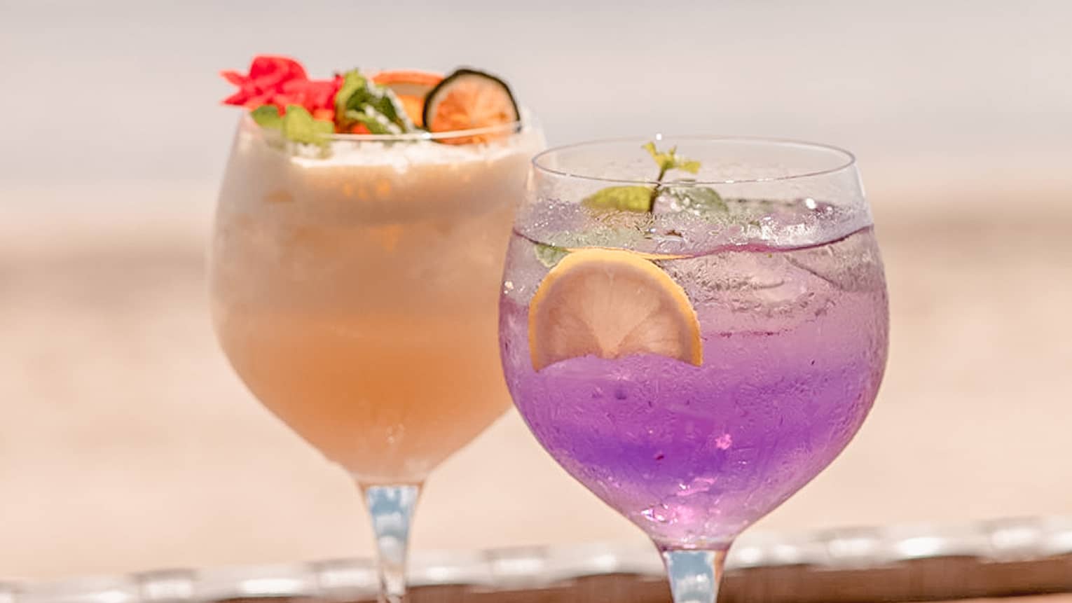Two colourful cocktails in stemmed glasses on wooden tray, beach in background