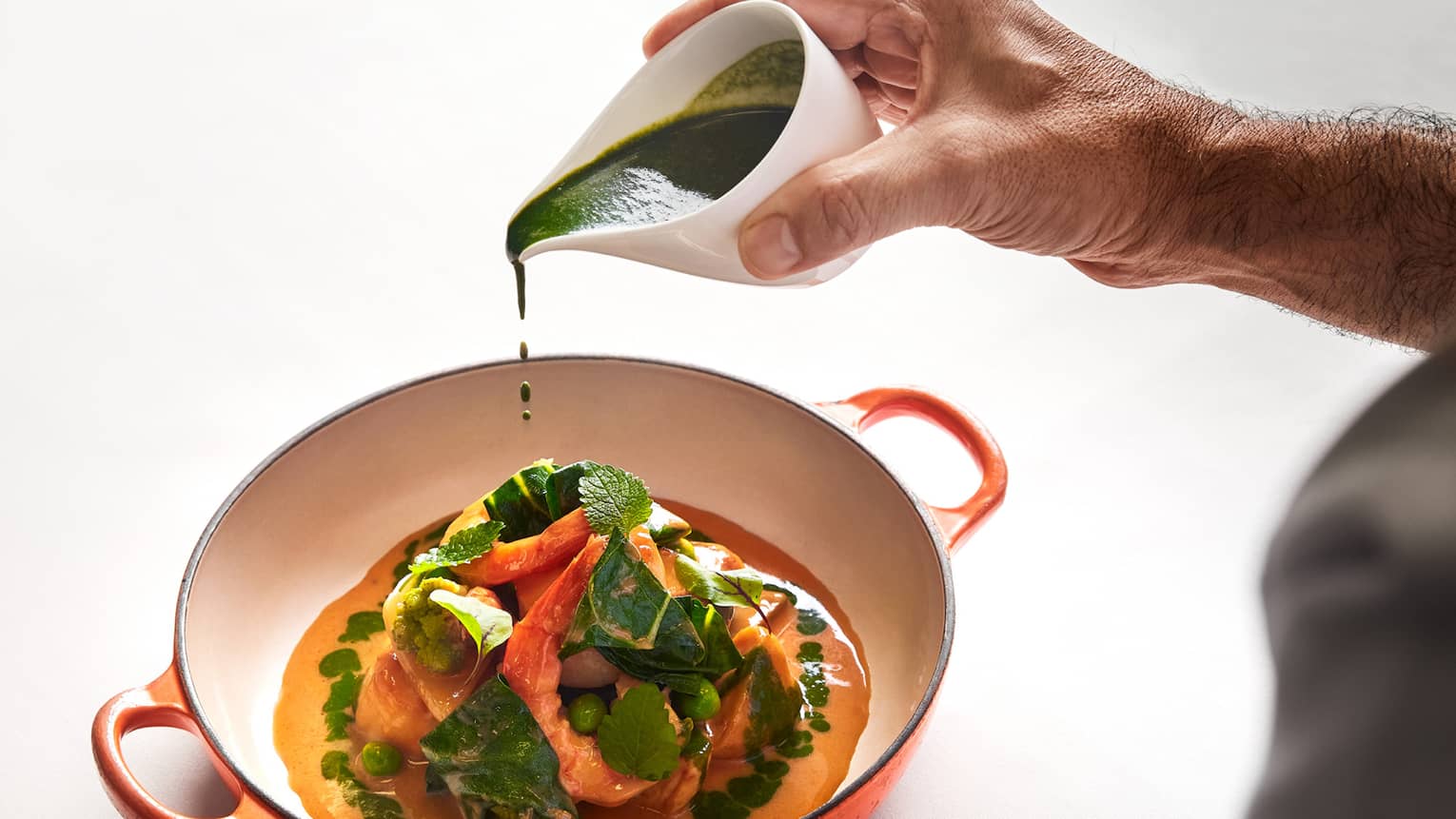Chef pouring green herb sauce over a vegetable and seafood dish in an orange ceramic bowl.