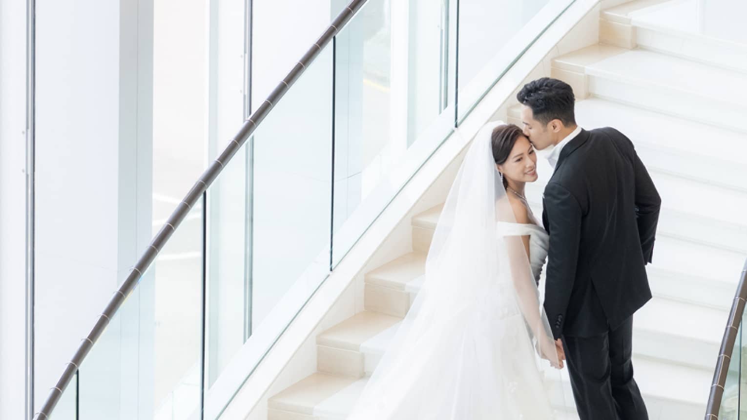 A bride and groom walking up stairs near large windows.