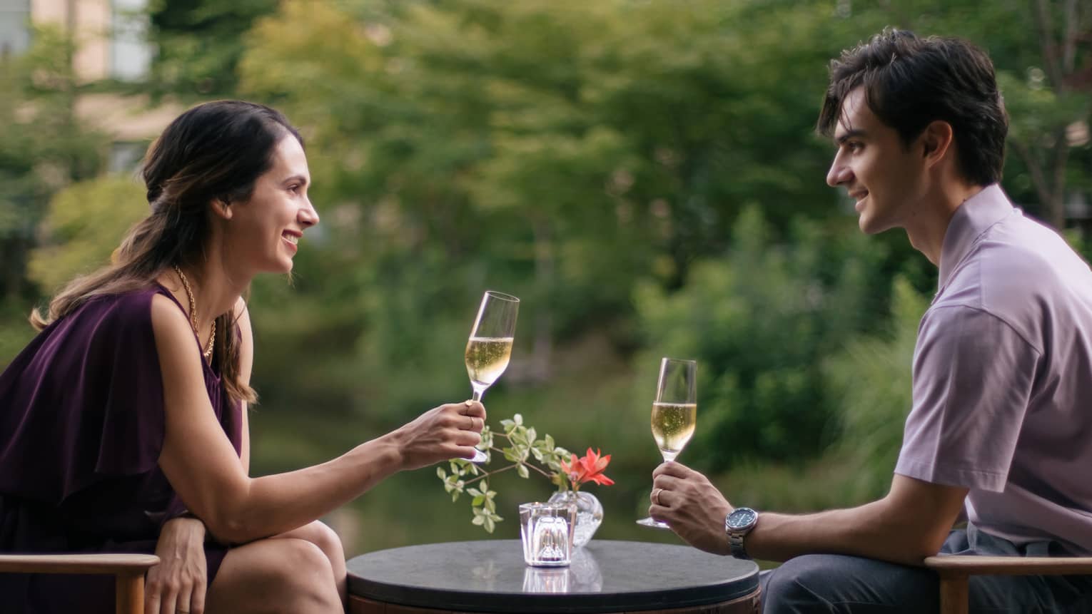 Two people sitting outdoors, toasting with champagne glasses at a small table with a flower vase