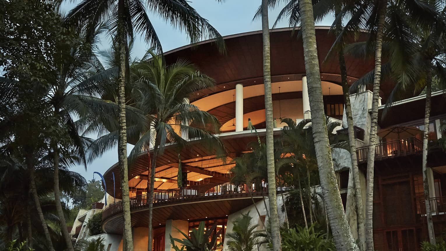 Four-storey curved balconies around resort building at dusk, lit with orange lights, surrounded by palms