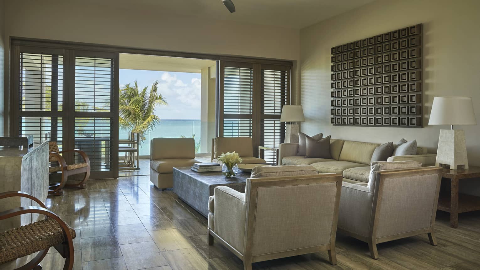 Bright living room with beige armchairs, sofa and ocean-view terrace framed by palm trees