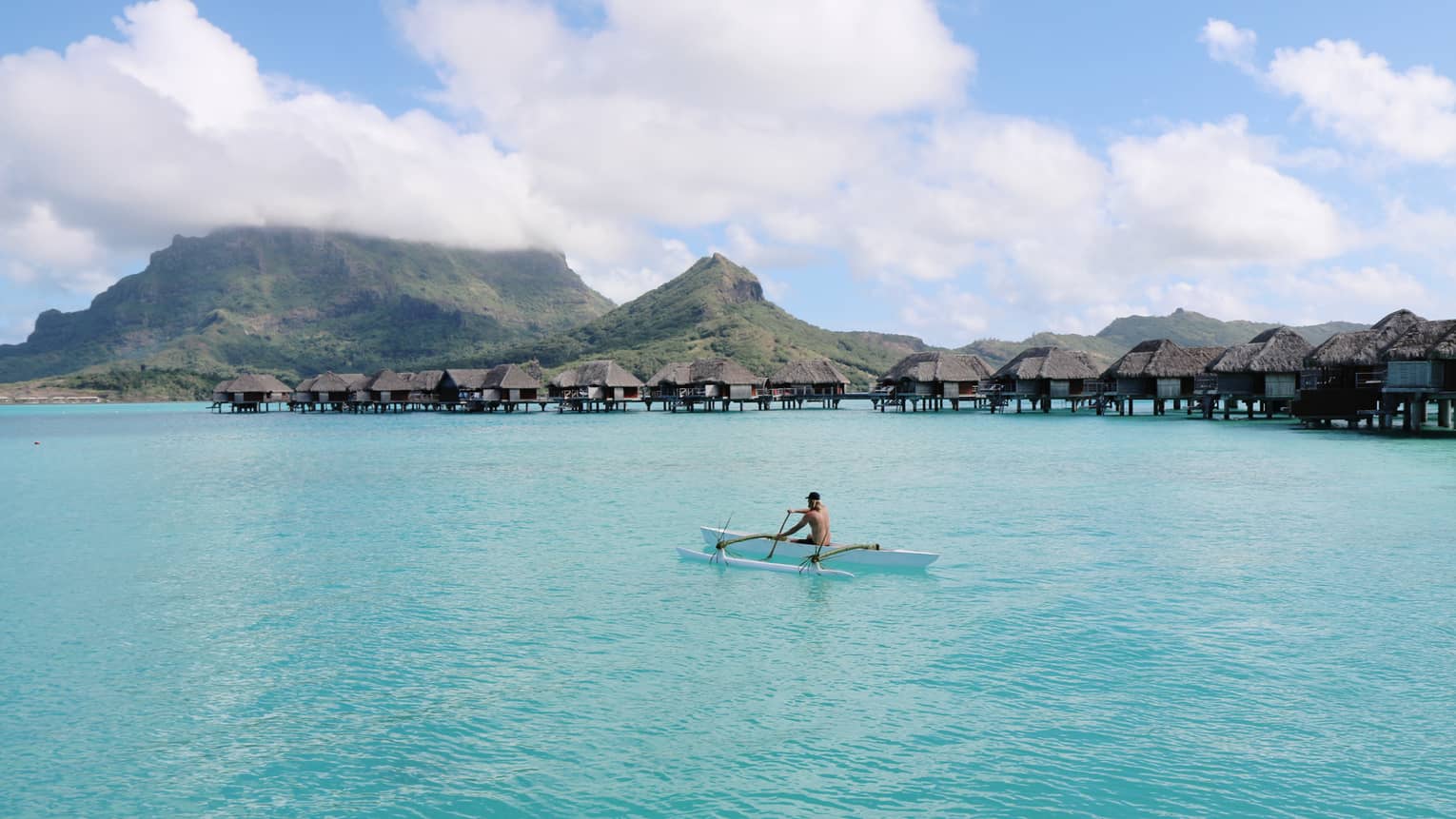 A guest paddles an outrigger canoe on clear turquoise water past a row of overwater bungalows and cloud-covered mountains.