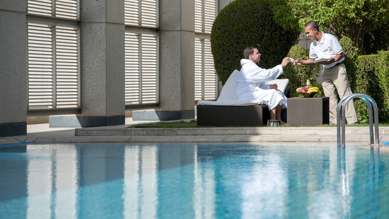 Man in white bathrobe sits on lounge chair on sunny swimming pool deck by fruit bowl, hotel stands hands him cloth