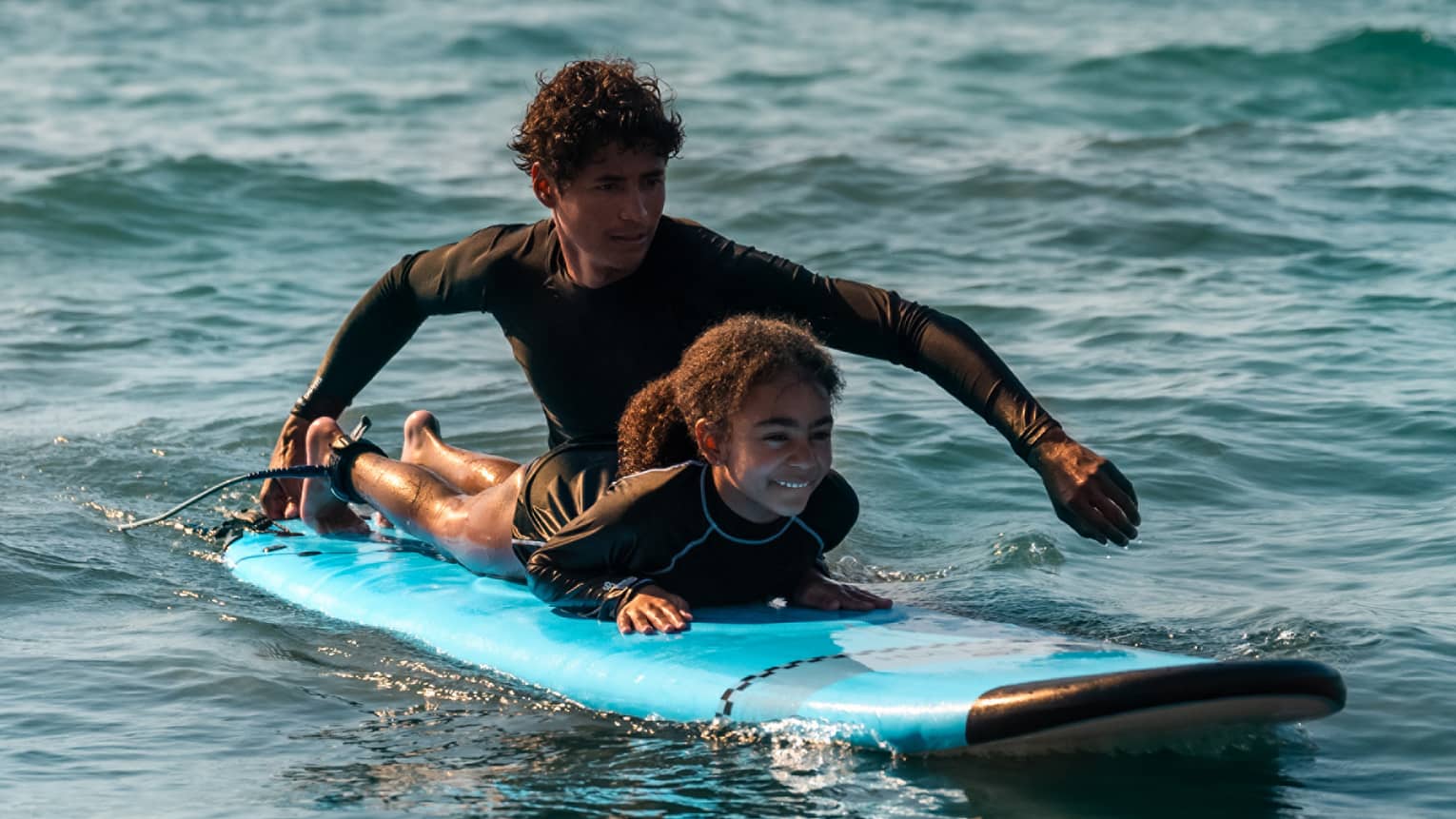 Male suf instructor wearing a black long-sleeve rashguard helps a young surfer paddle in the ocean