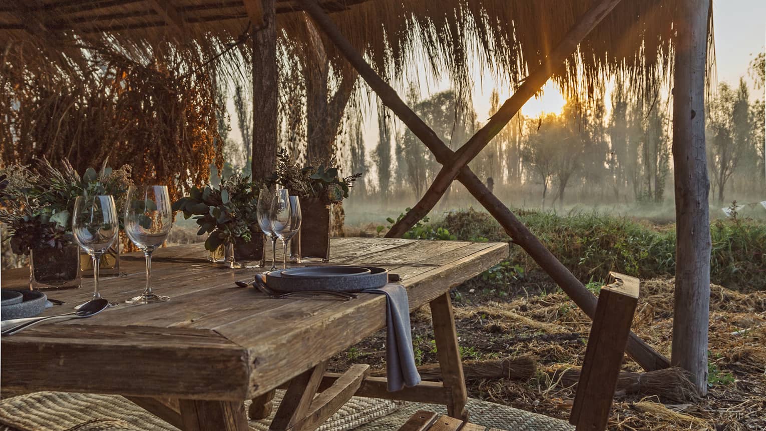 The setting sun shining on a thatched wooden canopy covering a wooden table with rustic dinnerware and succulent plants.