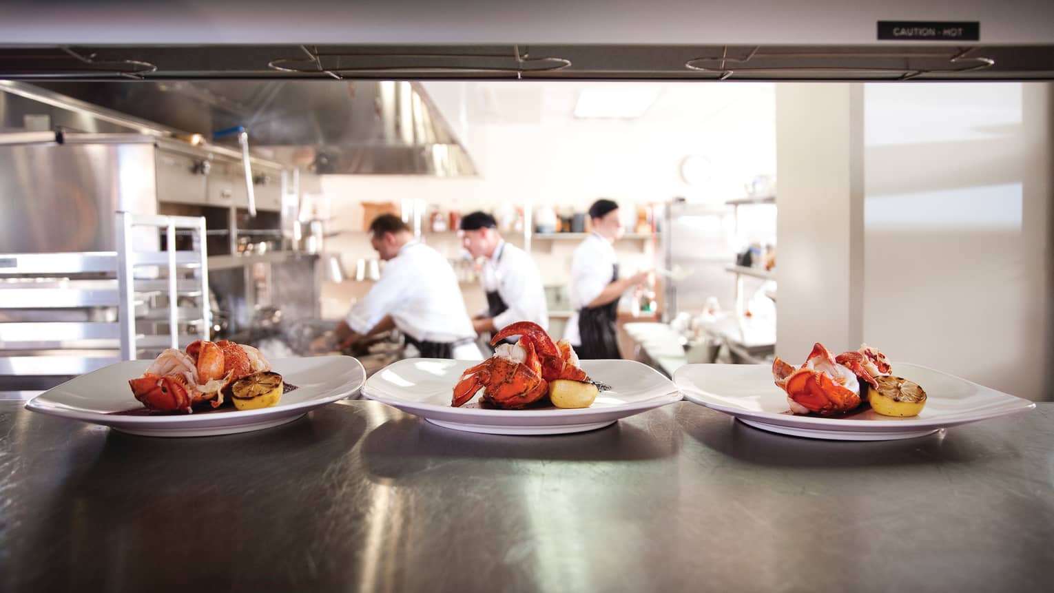 Three white plates with whole lobsters, lemons on kitchen lines, chefs in background