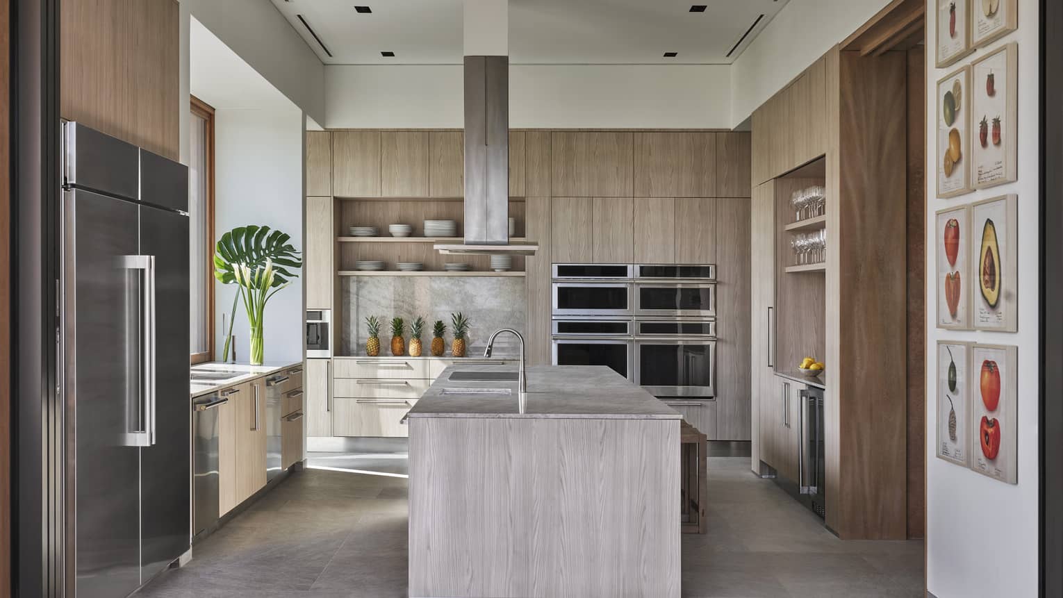 Kitchen with light wooden cabinetry, four ovens, island with hanging hutch, food artwork on the wall