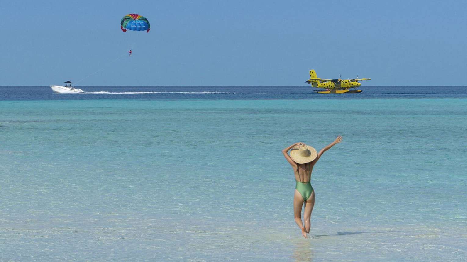 A woman walking on the beach watches a seaplane and a parasailor in the distance. 