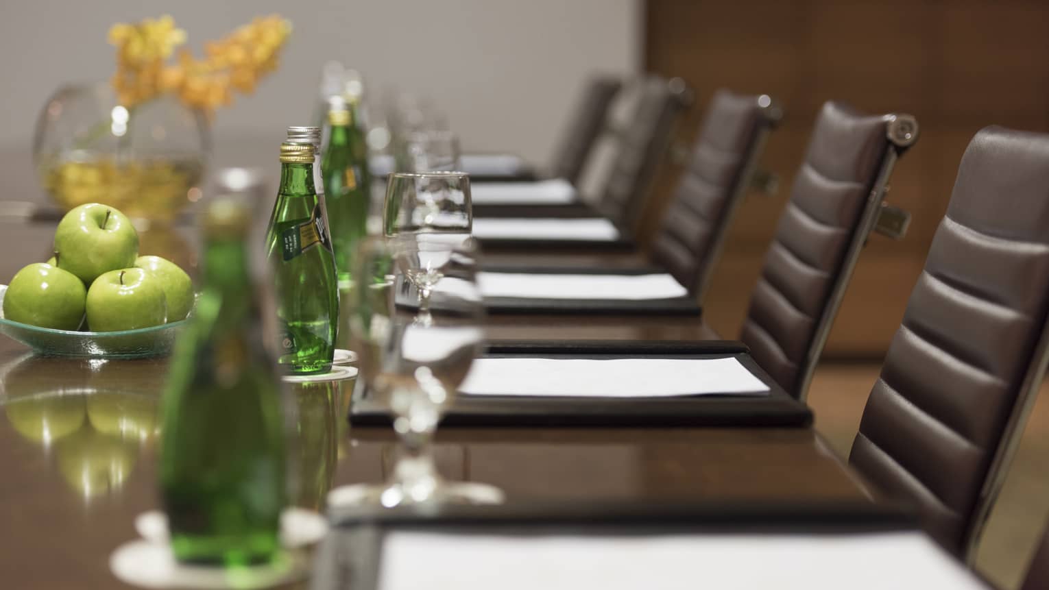 Close-up of business meeting table row of chairs, paper agendas, green glass bottles, apples