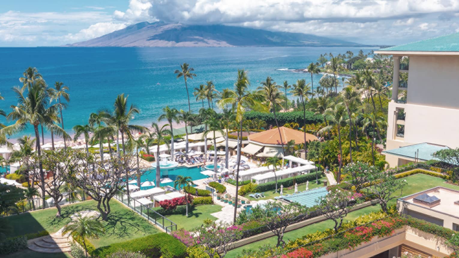Aerial view of resort with pools, palm trees, gardens and beach along a tropical coastline with mountains in the distance