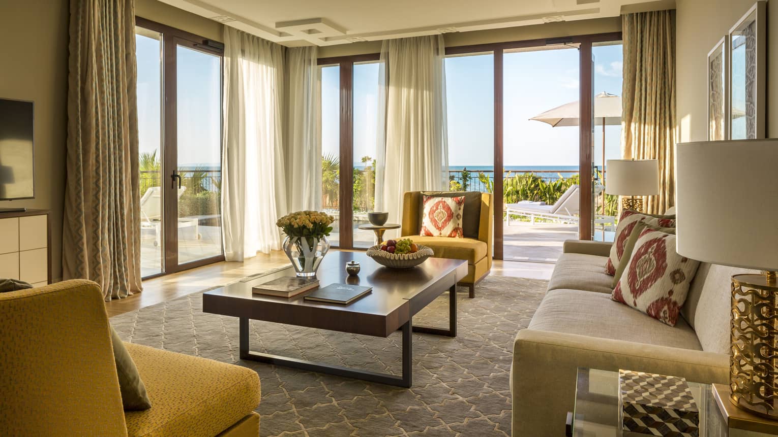 Living room with large rectangular table, beige sofa, mustard-yellow chairs, walls of windows looking out to sea