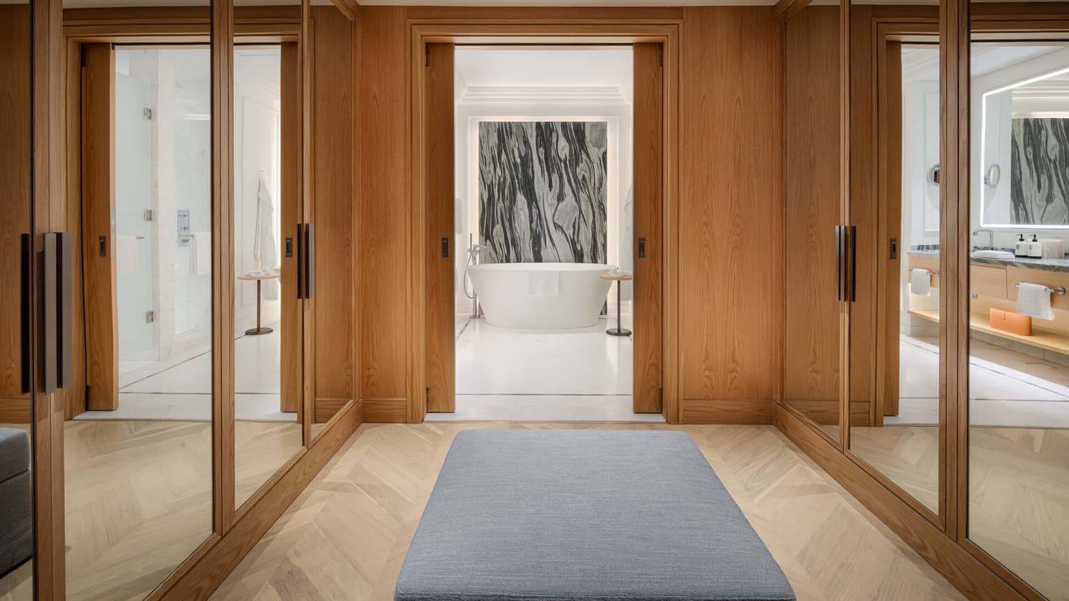 Modern walk-in closet with mirrored wooden wardrobes leading to a bathroom with a freestanding tub and marble accent wall.