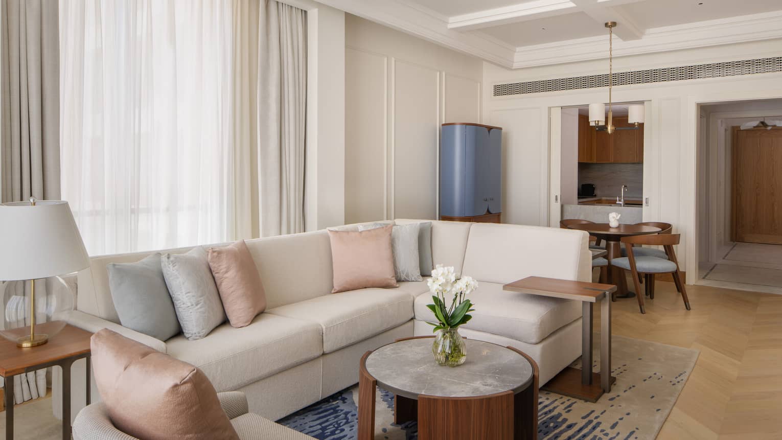 Modern living room with a cream sectional sofa, pastel cushions, round wood coffee table and dining area near an open kitchenette.
