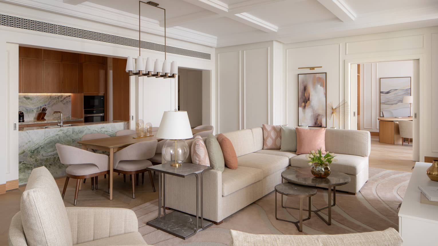 Elegant open-plan living space with neutral furnishings, pastel accents, a marble kitchen island and a dining area beside a modern chandelier.
