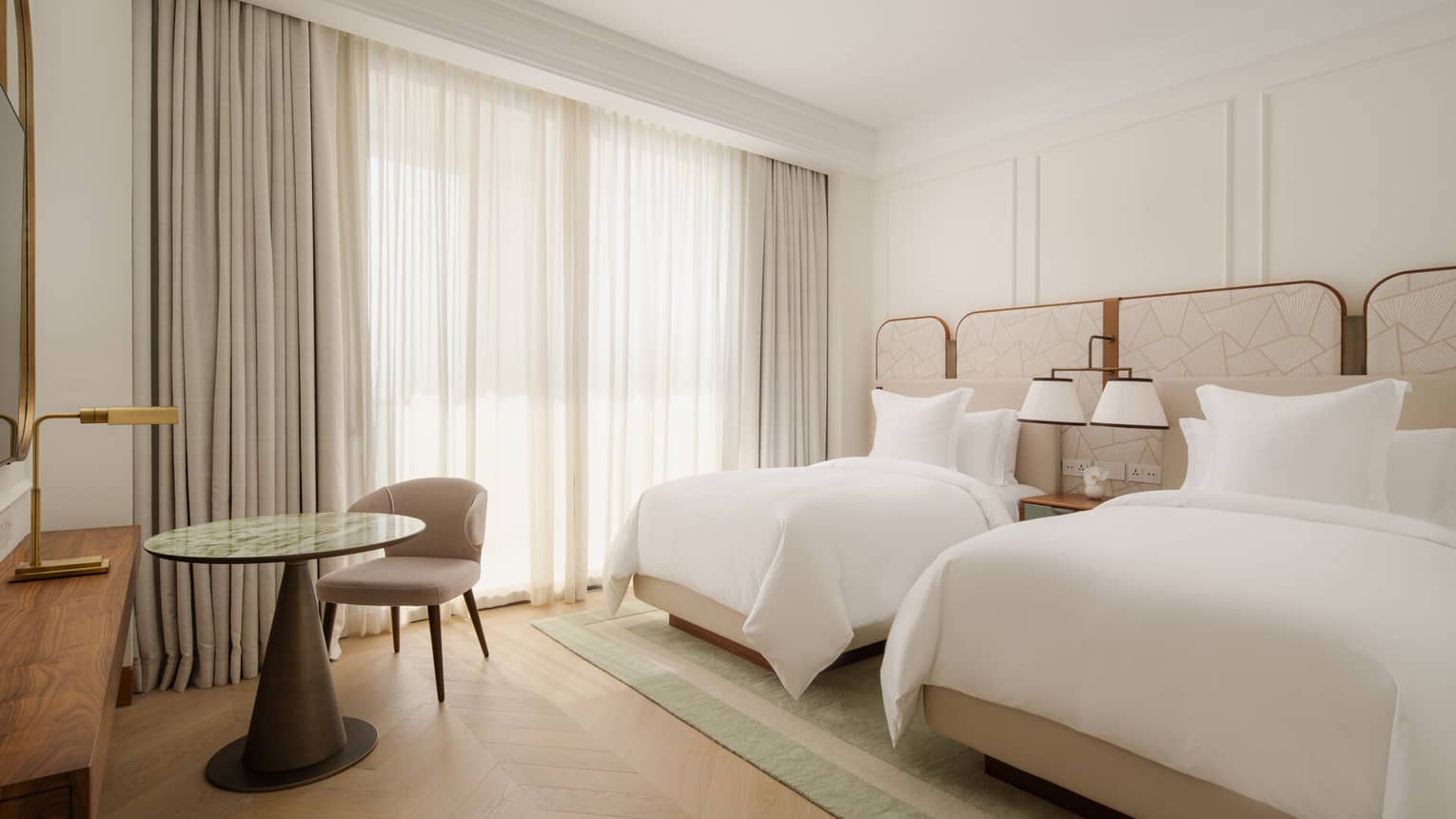 Bright bedroom with two single beds, neutral tones, a green rug, a small round table with a chair and floor-to-ceiling curtains.