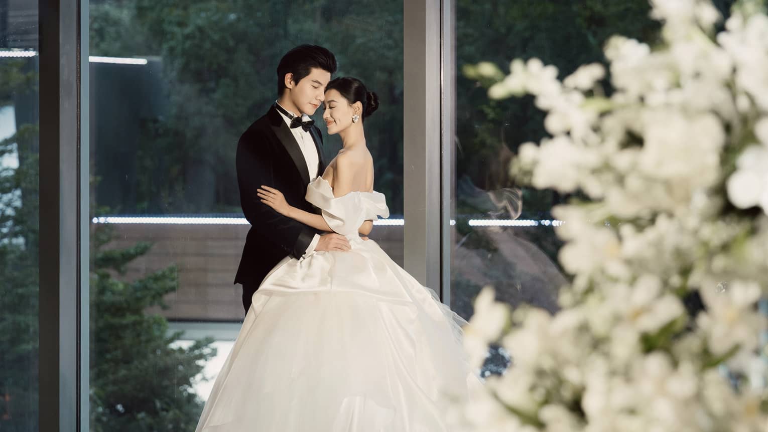 Bride and groom embrace in ballroom with floor-to-ceiling windows