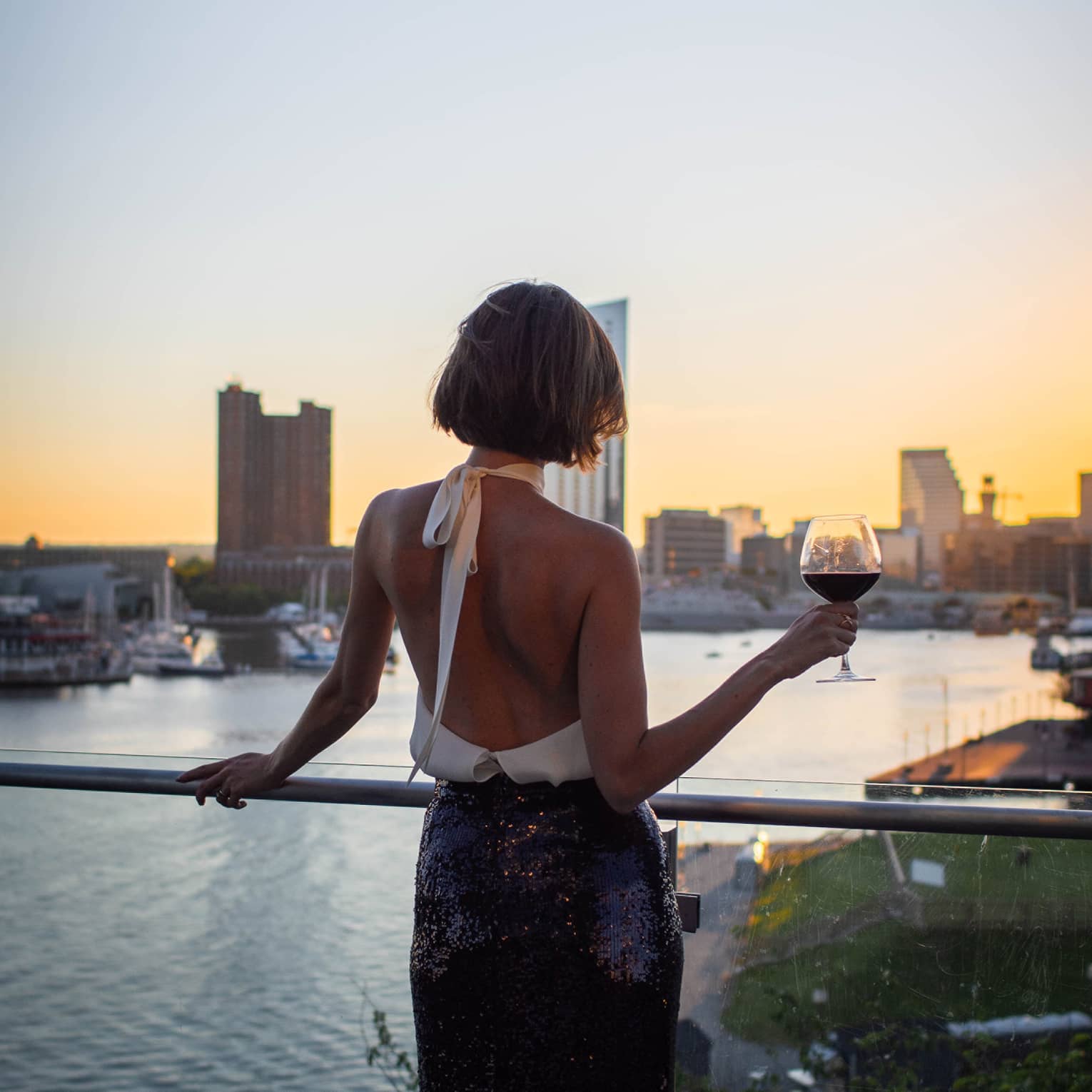 Person with bob haircut wearing a white halter-neck top and dark skirt stands silhouetted at sunset as they hold a glass of wine and overlook a city harbour