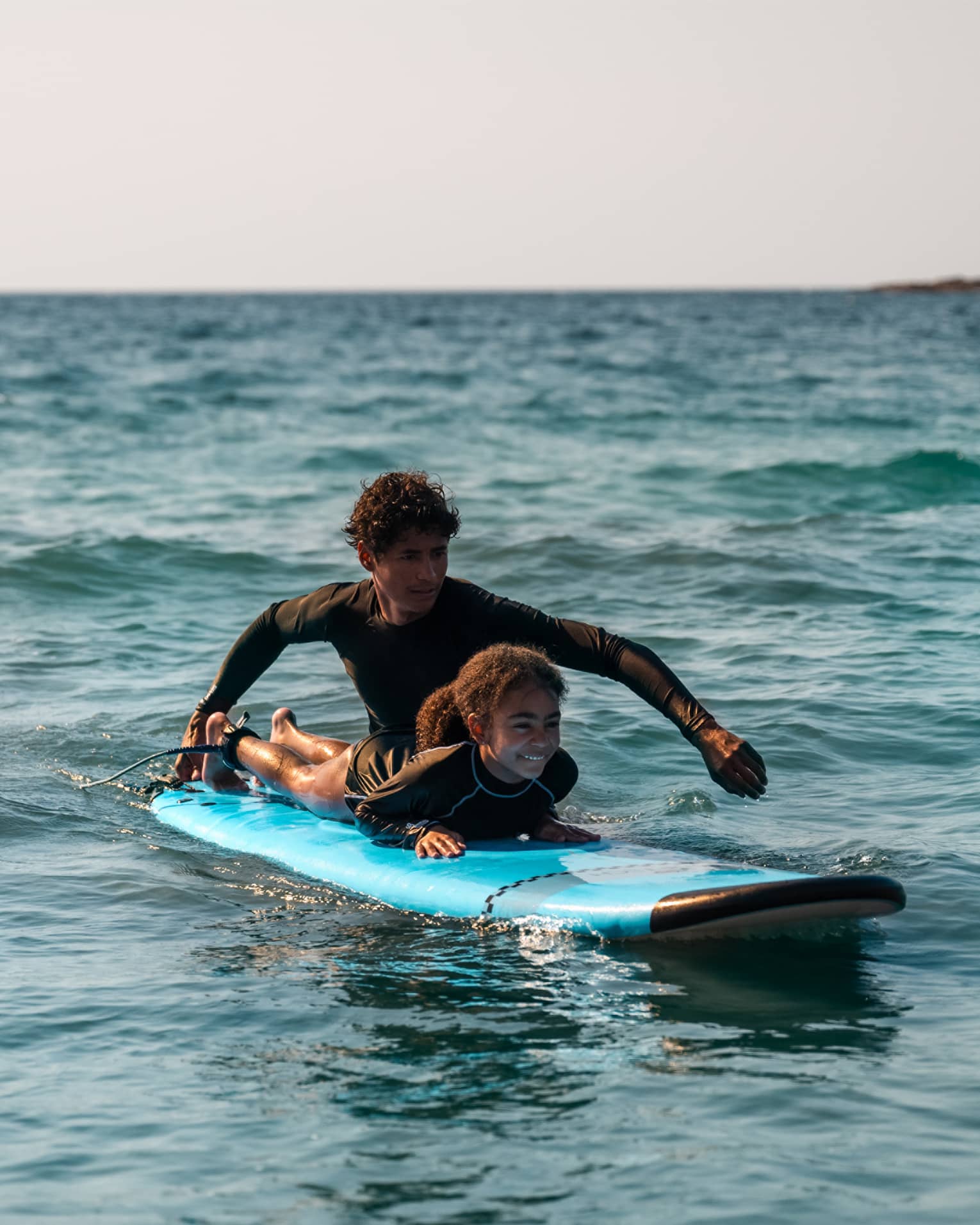 Male suf instructor wearing a black long-sleeve rashguard helps a young surfer paddle in the ocean