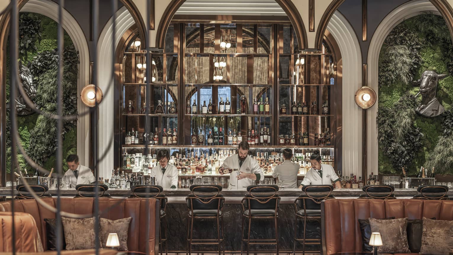 Chic bar with leather seating, plants on the walls and a well-stocked backlit bar, where bartenders in white jackets prepare drinks behind the counter.