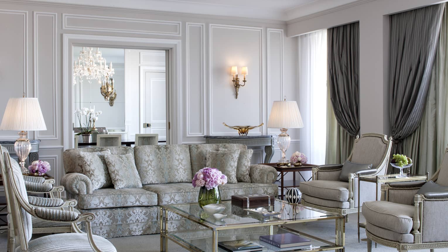Elegant living room with a patterned sofa, armchairs, glass coffee table, classic lamps and ornate wall paneling.