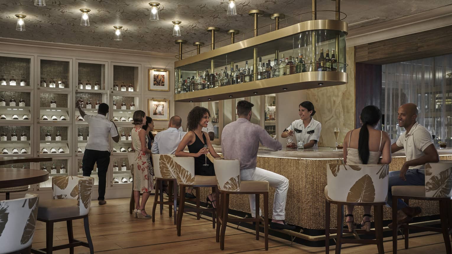 People mingle at a bar, with a hanging shelf holding bottles and a wall of wine shelving