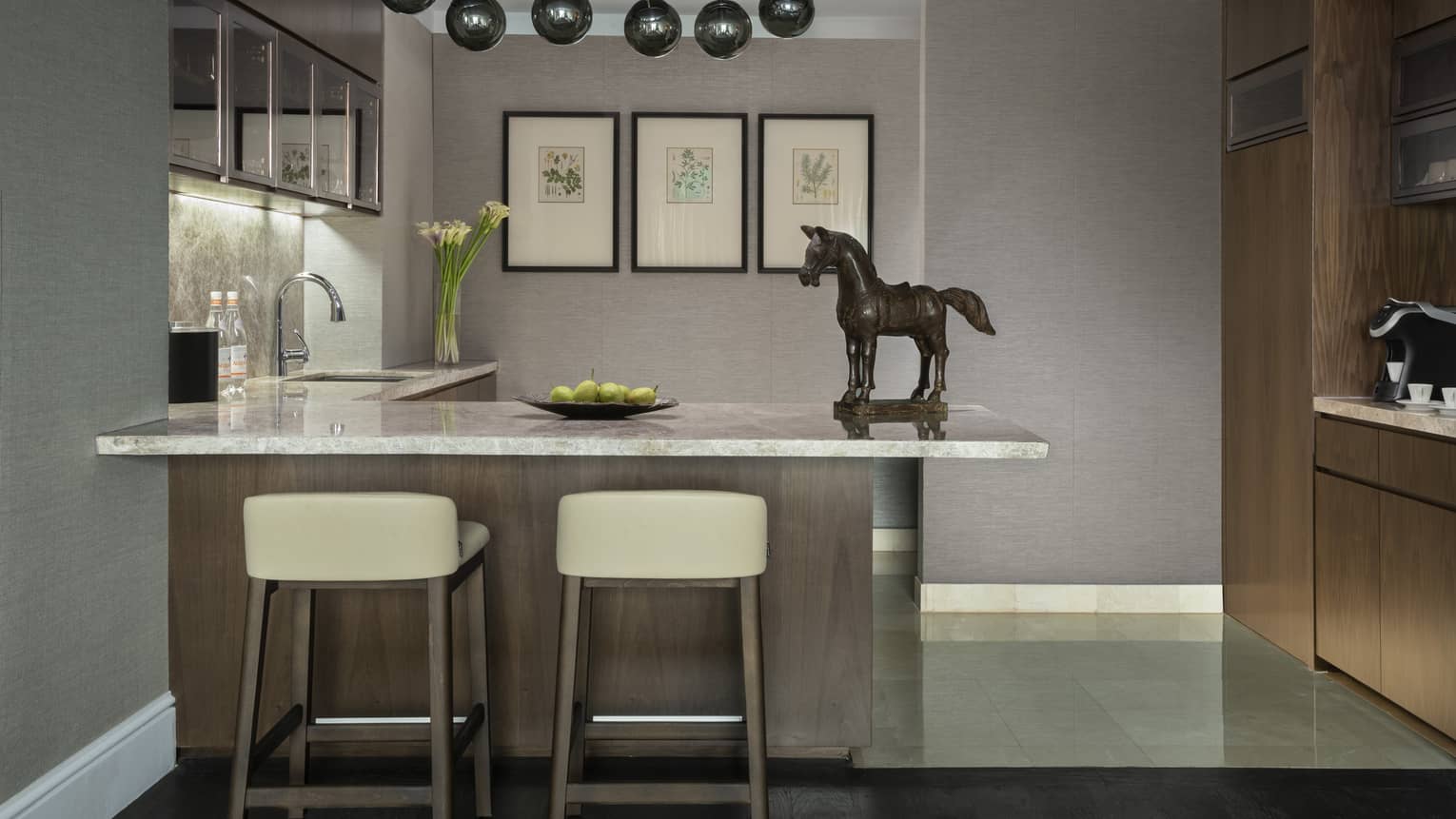 A modern kitchen area featuring a marble countertop island with two stools, decorative horse statue and framed botanical prints on the wall. The space includes warm wood accents and soft lighting.