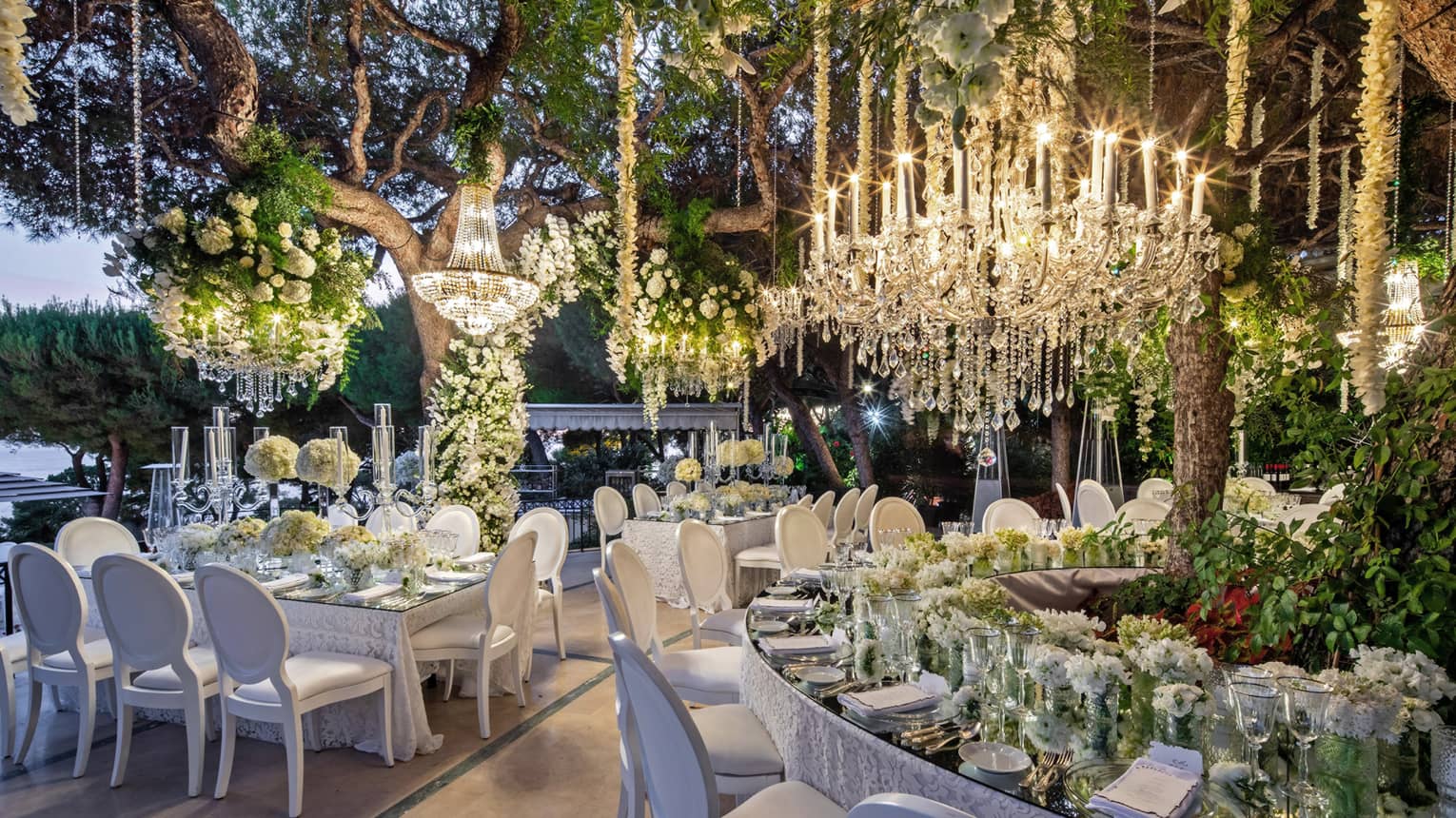 Elegantly set mirrored-top tables with white chairs, crystal chandeliers, floral arrangements hanging from ceiling