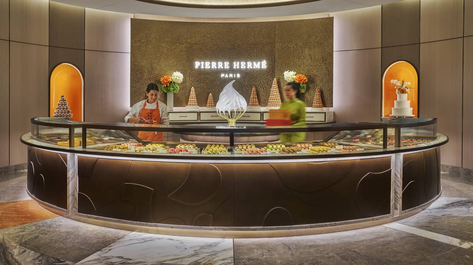 Person wearing white clothing and orange apron stands behind a round, glass-topped pastry counter
